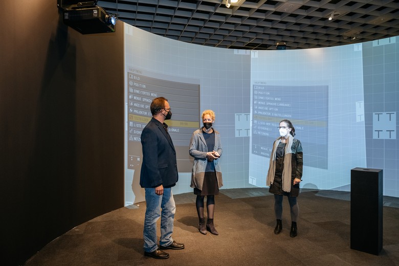Drei Personen stehen vor einer kahlen Wand, die Projektionen mit Menüverzeichnissen zeigt.