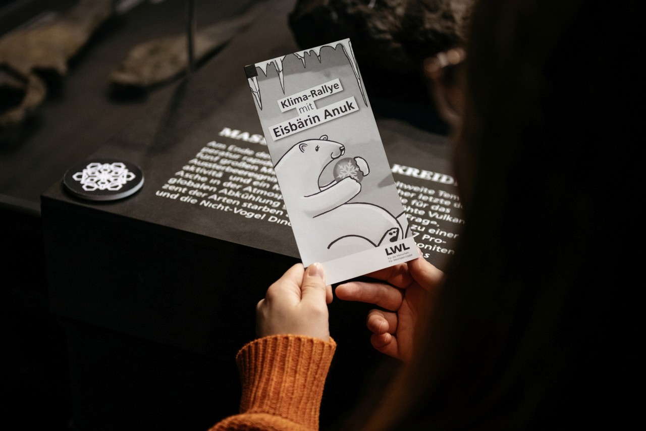 Eine Hand hält einen Handzettel auf dem ein Eisbär abgebildet ist. Im Hintergrund sieht man eine Infotafel mit Text und eine Schneeflocke.
