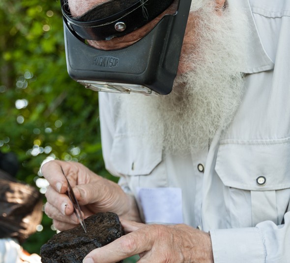 Ein bärtiger Mann mit Lupenbrille untersucht mit einer Pinzette ein Stück Moorboden.